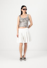 Ezüst flitteres crop top, fehér rakott szoknya és fényes cipők. A modell sötét napszemüveget visel, egy sima háttér előtt áll.