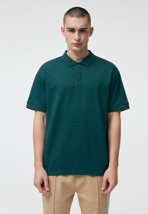 Młody mężczyzna z krótkimi włosami, ubrany w ciemnozieloną, wzorzystą koszulkę polo oraz beżowe spodnie, stojący na jednolitym białym tle.