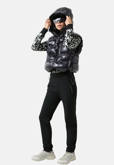 Kamizelka puchowa w błyszczącej czerni z kapturem, wzorzysta koszula z długim rękawem, czarne zwężane spodnie oraz jasnoszare trampki z grubymi podeszwami.