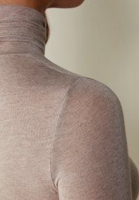 Dlouhý rukáv, přiléhavý rolák z měkké, světle hnědé pleteniny, s hladkým povrchem a bezešvým designem ramen.