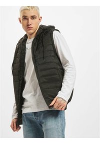 Zwarte puffer vest met een capuchon, featuring een doorgestikte textuur en een rits aan de voorkant. Gedragen over een wit langemouwen shirt en blauwe jeans.