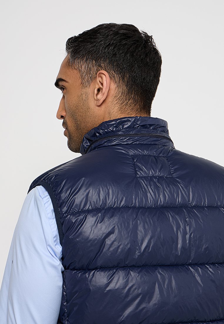 Gilet rembourré bleu marine avec col haut, texture matelassée et détails zippés. Matériau léger. Porté sur une chemise à manches longues bleu clair.