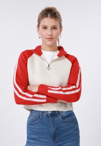 Κόκκινο και εκρού πουλόβερ με φερμουάρ και λευκές ρίγες στα μανίκια, διαθέτει ψηλό γιακά και στρογγυλό φερμουάρ. Συνδυάζε ται με μπλε τζιν.