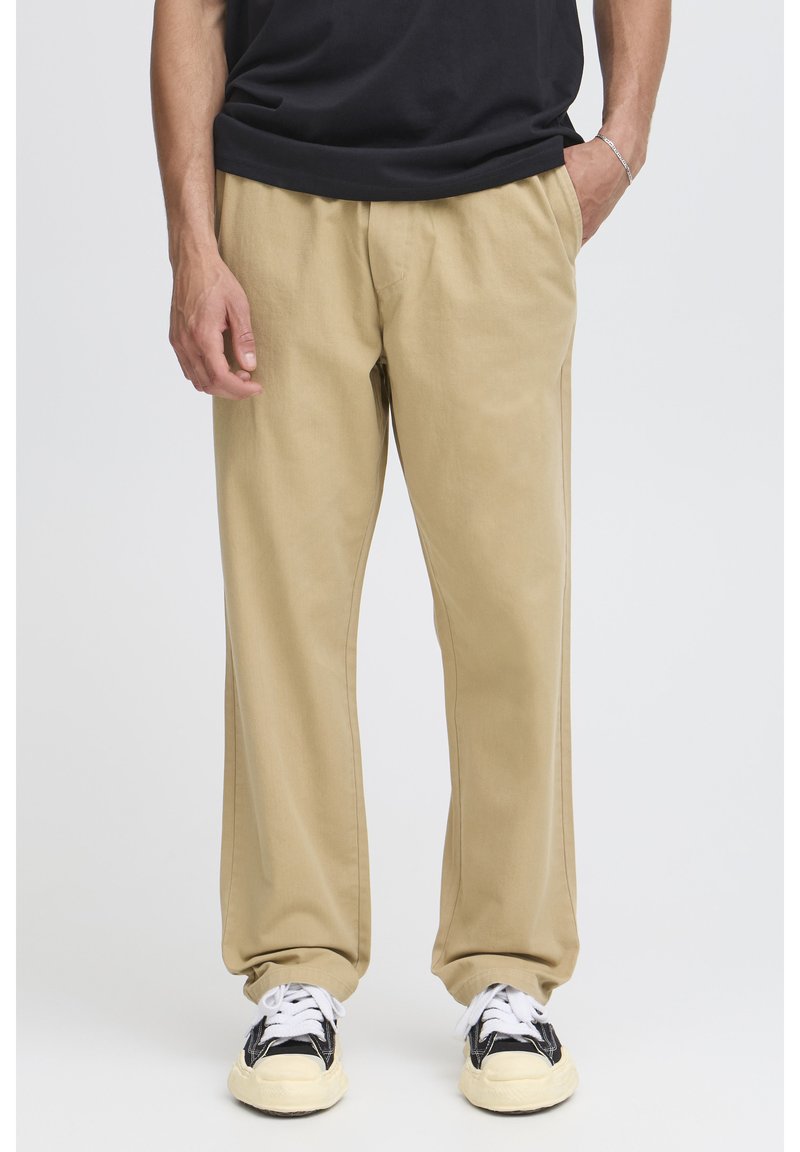 Pantaloni di cotone beige con vestibilità comoda, dotati di tasche laterali e una texture liscia. Indossati con sneakers nere e bianche.