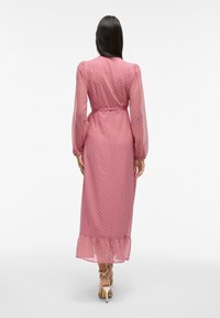 Abito lungo rosa con pois, maniche lunghe trasparenti, vita cintrata e orlo con volant. Indossato con scarpe con tacco alto. Vista posteriore.
