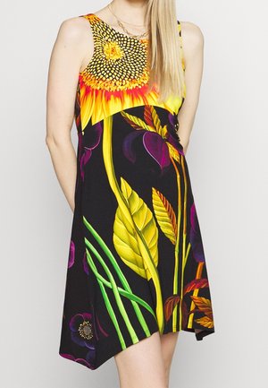 Hihaton mekko, jossa kirkas auringonkukka- ja monivärinen suuri kukkakuosi mustalla kankaalla, päällä pitkät vaaleat hiukset omaava henkilö.