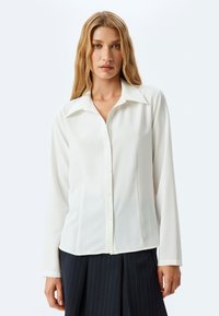 Blusa blanca de manga larga con cuello, botones en el frente y una tela suave y texturizada. Llevada con una falda oscura de rayas.