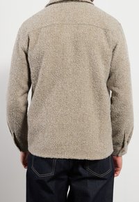 Beżowa, teksturowana kurtka o miękkim, futrzanym wykończeniu, z kołnierzem i zapinanymi mankietami, zestawiona z ciemnoniebieskimi dżinsami.