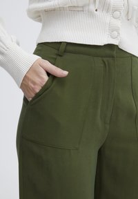Pantaloni verdi realizzati con un tessuto morbido, dotati di tasche laterali e dettagli a pieghe. Indossati con un maglione bianco in maglia.