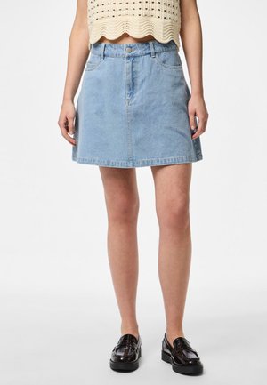 Donna che indossa una minigonna di jeans azzurro chiaro, un top corto beige all'uncinetto e mocassini marrone scuro, in piedi davanti a uno sfondo semplice.