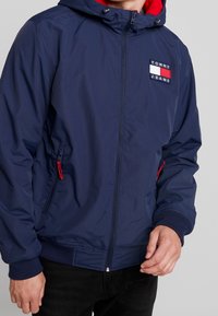 Marineblaue Außenjacke mit roter Kapuze, Reißverschluss vorne, Seitentaschen und gesticktem "TOMMY JEANS"-Logo-Aufnäher auf der Brust.