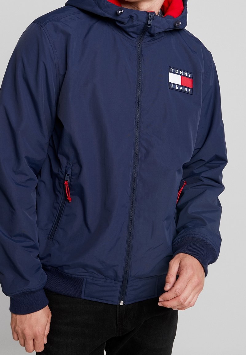 Marineblaue Außenjacke mit roter Kapuze, Reißverschluss vorne, Seitentaschen und gesticktem "TOMMY JEANS"-Logo-Aufnäher auf der Brust.