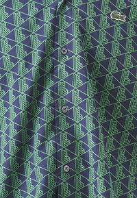 Cămașă cu mânecă scurtă de culoare albastru închis, cu un model geometric verde. Are un sistem de închidere cu nasturi pe față și un logo mic pe piept.
