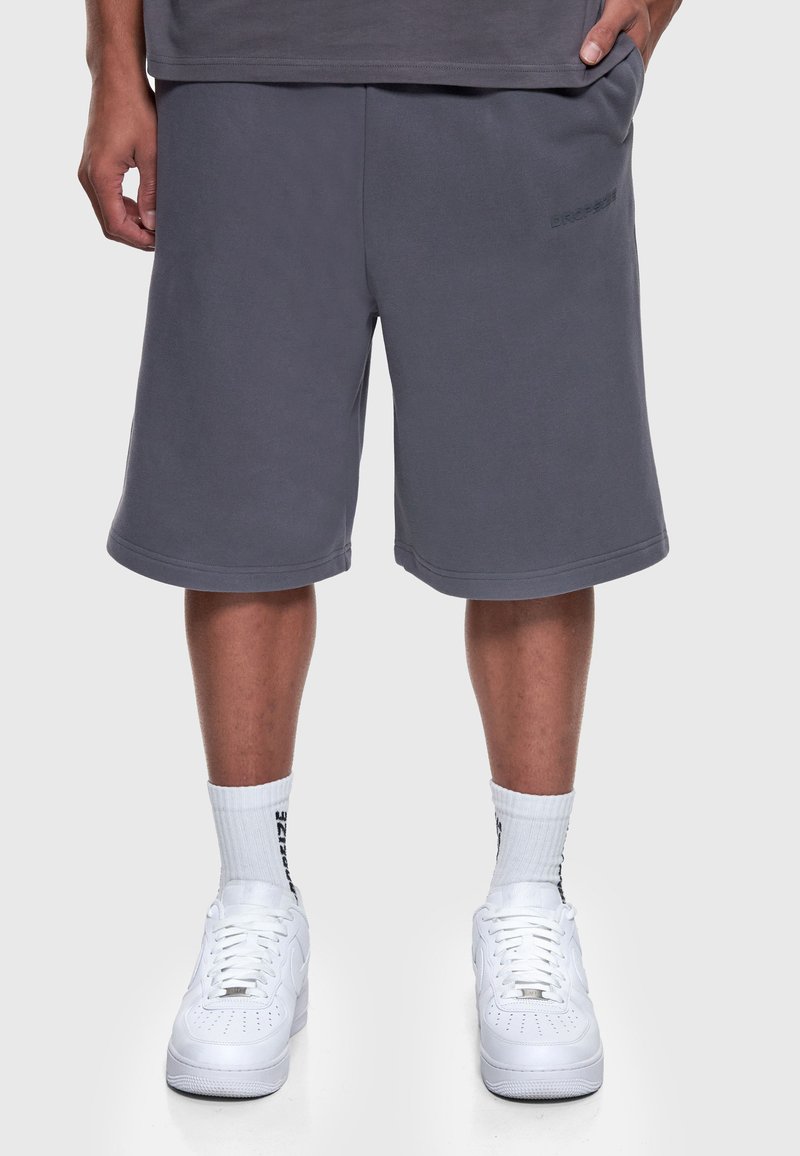 Homme portant un short gris jusqu’aux genoux, des chaussettes blanches avec un texte noir, et des baskets blanches, debout, les mains relâchées le long du corps.