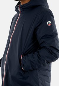 Giacca blu navy con cappuccio, dotata di chiusura con zip rossa, polsini elasticizzati e tasche laterali. Applicazione del logo nella parte superiore del braccio. Materiale leggero.