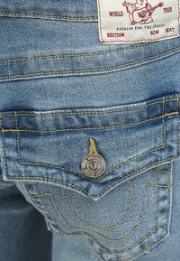 Denim jeans met blauwe stof, gele stiksels en een zilveren knop met branding. De achterzak heeft een gebogen stiksel en label.