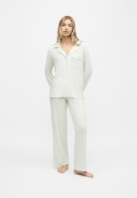 Femme aux longs cheveux blonds, debout pieds nus, portant un haut gris clair à manches longues avec boutons et un pantalon ample assorti sur fond blanc.
