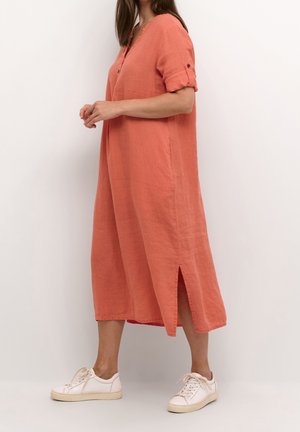 Vrouw in een losvallende koraalkleurige jurk van middelmatige lengte met opgerolde mouwen en een zijsplit, gekleed in witte sneakers, staand tegen een effen lichte achtergrond.