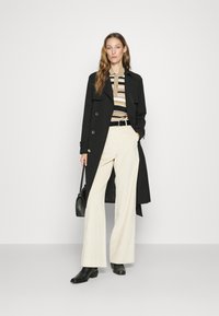 Svart trenchcoat med gullknapper, rutete beige og svart skjorte, hvite vide bukser, svart belte, og ankelstøvler. Liten svart håndveske.