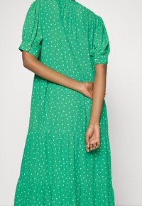Robe verte à pois blancs, manches courtes bouffantes, design à volants, et encolure froncée. Le tissu semble léger et texturé.