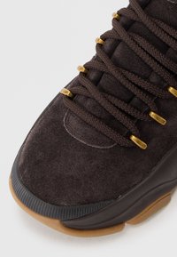 Brown suede sneaker featuring textured surface, gold eyelets, thick brown laces, and a rubber sole with a slight grip pattern.