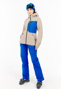 Lichtbeige skijas met blauwe accenten, ritszakken en capuchon; gecombineerd met blauwe skibroeken en zwarte skibotten.