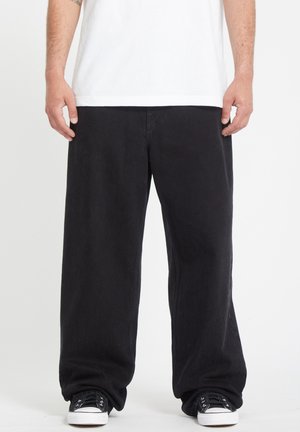 Homme portant un pantalon noir ample, des baskets blanches et un t-shirt blanc uni, debout devant un fond blanc uni.