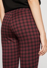 Bordowe legginsy w kratkę o miękkiej fakturze, z czarnym wzorem siatki i elastycznym pasem. Zbliżenie z tyłu.