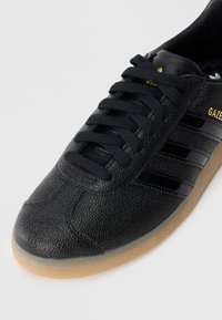 Sort lædersneaker med en tekstureret finish, der har sorte snørebånd, tre sorte striber og en gummisål. Guld "GAZELLE" branding.