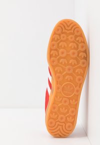 Röd sneakersko med vita ränder, texturerad mockaovandel och gummisula med ett cirkulärt mönster samt Adidas-logotyp på hälen.