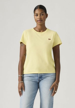 Mujer con el pelo trenzado, vestida con una camiseta amarilla de Levi’s y vaqueros azul claro, de pie frente a un fondo claro y sencillo.