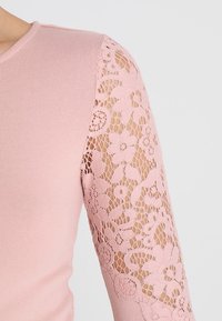 Vaaleanpunainen pitkähihainen paita, jossa on rohkeasti strukturoidut ribbidet ja herkkä kukkakuvioinen pitsi yhdellä hihalla, jonka viimeistely on läpikuultava.