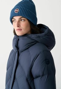 Blå stickad mössa och en huva pufferjacka i mörknavy. Jackan har en subtil lyster, quiltat mönster och logodetalj på ärmen.