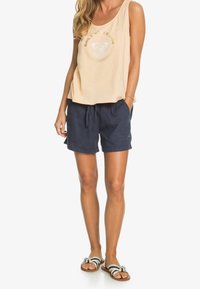 Vrouw in een beige mouwloos top met logo, marineblauwe shorts met trekkoord en gestreepte sandalen, staand tegen een witte achtergrond.
