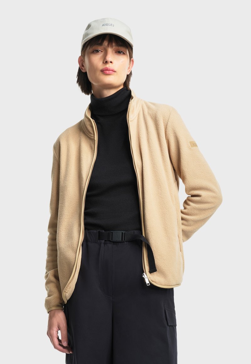 Veste en polaire beige avec un col montant et une fermeture éclair sur le devant, portée sur un pull à col roulé noir. Comprend des poches latérales et un design simple.