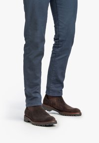 Stivali alla caviglia in suede marrone con pannelli laterali elastici e suole in gomma robuste, abbinati a pantaloni aderenti blu navy.