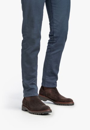 Bruine suède enkelboots met elastische zijpanelen en robuuste rubberen zolen, gecombineerd met strakke marineblauwe broek.