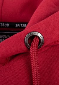 Rode hoodie stof met een gladde textuur, voorzien van een zilveren metalen oogje en een gevlochten koord. "SPITZBUB" branding zichtbaar op het label.