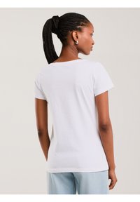 Naine punutud hobusesabaga, kes kannab hõbedast rõngakujulist kõrvarõngast, lihtsat valget lühikest varrukat särki ja hele siniseid teksapükse, selg kaamerale pööratud.