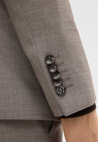 Hellgrauer, texturierter Blazerärmel mit fünf dunklen Knöpfen, der eine Mischung aus glatten und strukturierten Oberflächen sowie einem schwarzen Strickbundakzent aufweist.