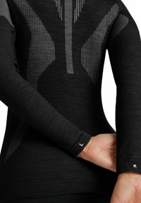 Sous-couche à manches longues noire avec un motif texturé, des accents gris clair, et des étiquettes claires 'L' et 'R' sur les poignets pour le taillage.