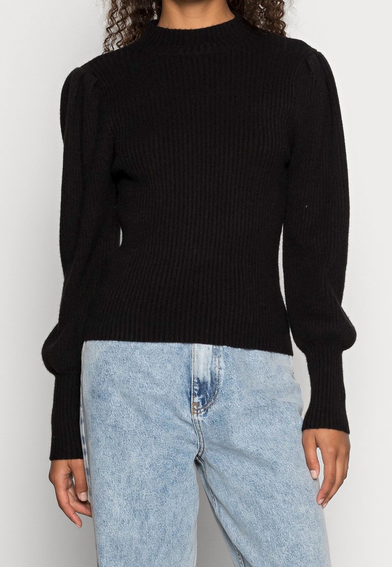 Zwarte geribde gebreide trui met een hoge hals, aansluitend lijfje en opgeblazen lange mouwen, gecombineerd met lichtblauwe high-waisted jeans.