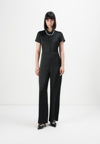 Woman standing against white background wearing a black short-sleeve jumpsuit, pearl necklace, and black pointed heels with ankle straps.