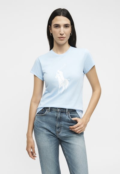 Γυναίκα με μακριά σκούρα μαλλιά, που φοράει ανοιχτό μπλε μπλουζάκι πόλο με λογότυπο και μπλε τζιν, στέκεται με το ένα χέρι στην τσέπη μπροστά σε άσπρο φόντο.