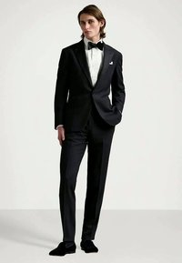 Homem a usar um smoking preto com uma camisa branca e uma gravata borboleta preta, em pé com uma mão no bolso contra um fundo simples.