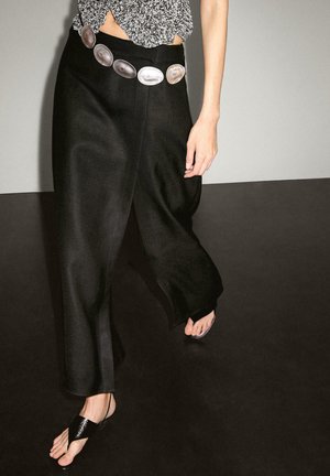 Donna che indossa pantaloni larghi neri con una cintura a grande pietra ovale e sandali neri con punta aperta, in piedi su uno sfondo con pavimento e parete bicolore.