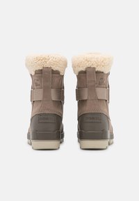 Een paar bruine winterlaarzen met beige rubberen zolen, verstelbare banden en een pluizige crème-kleurige fleece voering die zichtbaar is aan de bovenkant, gezien van achteren.