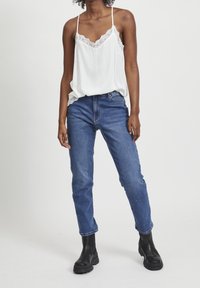 Caraco blanc orné de dentelle, associé à un jean bleu taille haute. Le modèle porte des bottines noires épaisses. Design simple et décontracté.