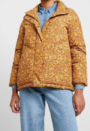 Person trägt eine senfgelbe gesteppte Jacke mit Blumenmuster über einem hellblauen Jeanshemd und Jeans, steht mit locker hängenden Armen an den Seiten.