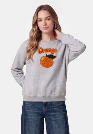 Ung kvinde med langt blondt hår bærer en grå sweatshirt med en appelsinfrugt og teksten "Orange", kombineret med blå jeans og blå øreringe i hoop-stil.